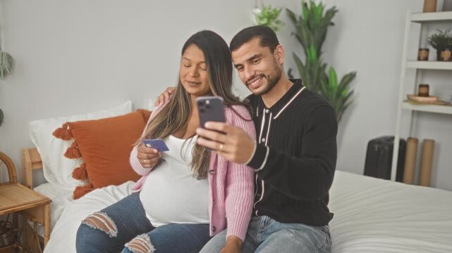 Man holding smartphone takes selfie with pregnant woman showing belly and creditcard seated on bed in a studio; expectant family joy.