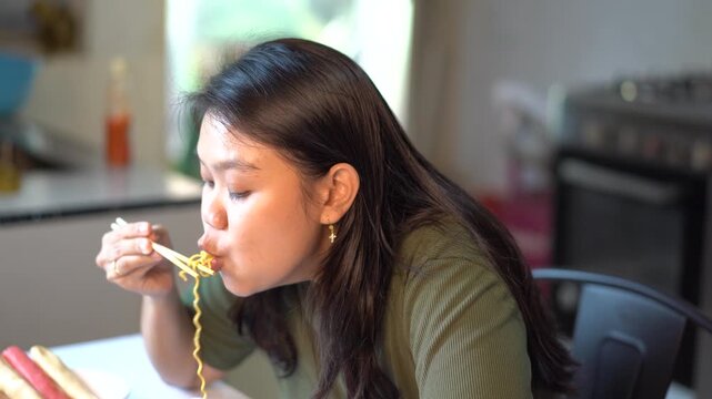 Asian woman eating instant noodles with chopsticks in the kitchen