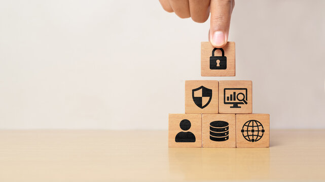 Hand placing lock icon wooden block on cybersecurity pyramid with user, database, network, shield and monitoring symbols, representing data protection, digital security and risk management.