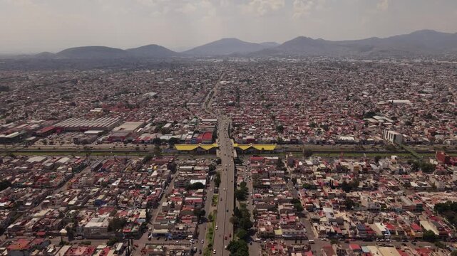 Aerial drone view approaching Avenida Central and Metro Line B in Ecatepec, Mexico, with cars, pedestrians, buildings, mountains and white clouds on a sunny day
