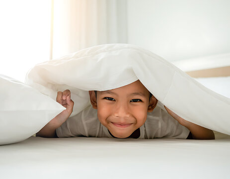 Happy Little Asian Boy Hiding Under White Blanket in Bright Bedroom Looking at Camera with Smile