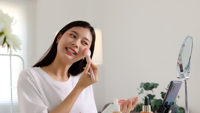 Young Asian beauty influencer applying blush with a powder brush while recording a makeup tutorial on smartphone in her room, perfect for cosmetics, blogging, and social media content.