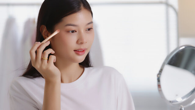 Close-up of a young Asian woman applying eyeshadow with a small makeup brush in front of a mirror, ideal for cosmetics, beauty tutorials, and skincare content.