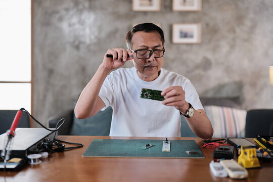 Mature Asian man looks at and considers circuit board details with magnifying glass, focusing on precision checks for home electrical appliance DIY fix, electronics component hobby, handyman workshop.