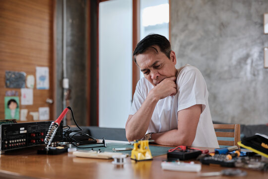 Mature Asian man concentrates on repairing home electrical appliance, disassembling and soldering circuit board, tired from precision detail focusing, taking a break, resting from his workshop hobby.