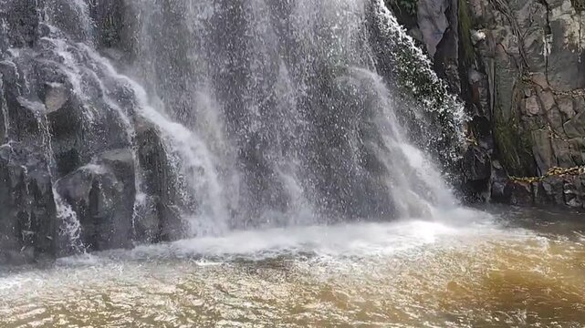 A scenic and powerful waterfall cascades down dark rocky cliffs into a turbulent pool of water in a serene natural forest landscape