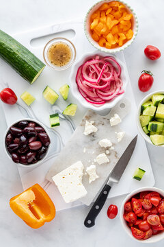 A white marble cutting board topped with ingredients for making a Greek salad.