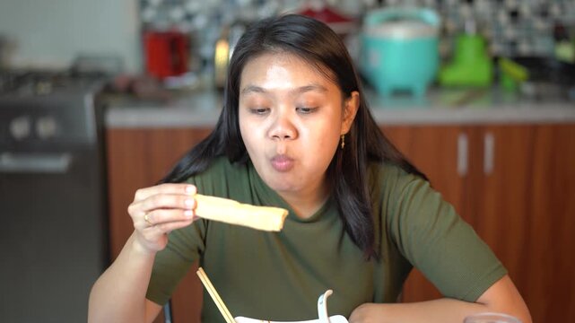 Asian woman eating a spring roll with chopsticks in the kitchen