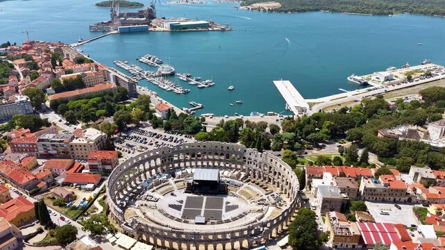 Aerial drone view of the Roman amphitheatre with a stage inside it, the harbour and the boats behind, Pula, Croatia