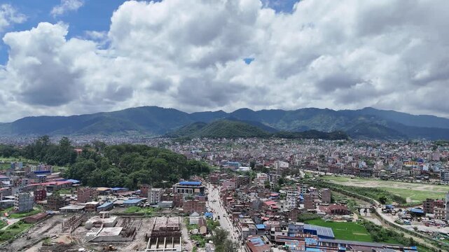 Aerial view of a dense Nepalese city featuring a prominent forested hill surrounded by urban sprawl and distant mountain ranges
