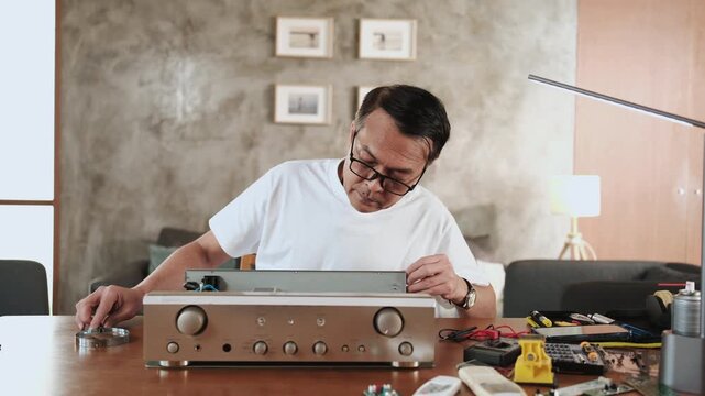 Mature Asian man disassembling and checking audio equipment, looking with magnifying glass at circuit board details for repair, home appliance DIY fix, electronics component hobby, handyman workshop.