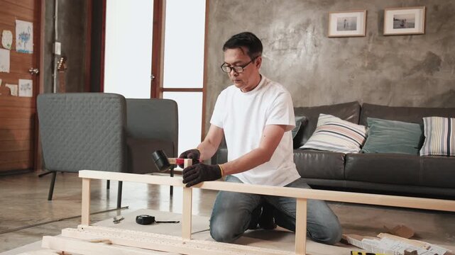 Asian mature man self-assembling furniture with DIY tools, nailing joint parts with rubber mallet, checking precision, home improvement, craft installation, hobby wooden workshop, active lifestyle.