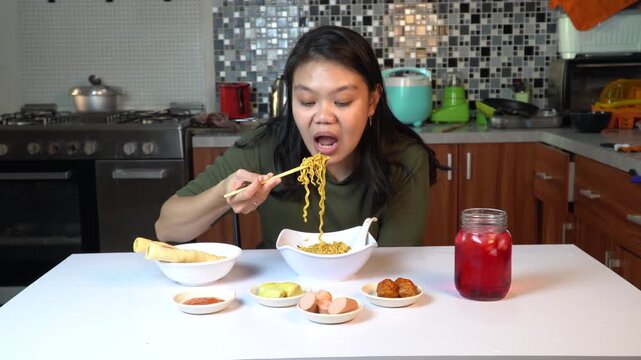 Asian woman eating instant noodles with chopsticks in kitchen at home