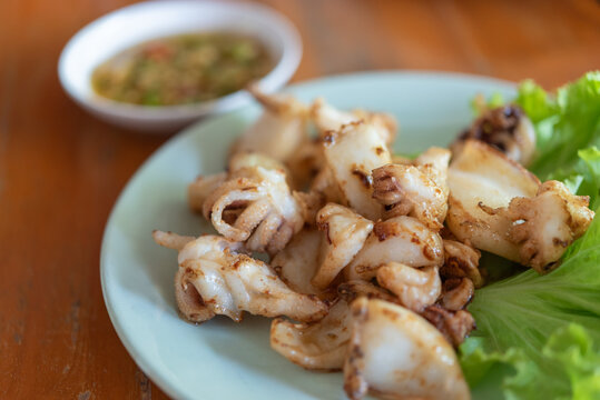Grilled squid pieces served fresh green lettuce spicy seafood dipping sauce plate offering savory Thai meal tender texture smoky flavor