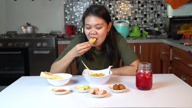 Young Asian woman eating spring rolls and noodles in her kitchen.