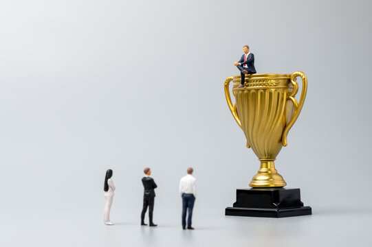Miniature businessman sitting atop a giant gold trophy while a group looks up from the ground. Social class, economic disparity, corporate hierarchy concept.