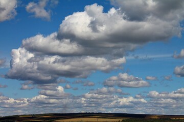 Soft white clouds drift across a clear blue sky above distant fields in green, yellow, and terracotta tones, creating a peaceful rural landscape with an open, balanced feel. © Liudmila