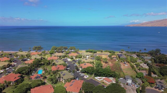 Aerial view over Kihei on Maui Hawaii island showing red tile roof condominiums palm trees swimming pools parked cars a sandy shoreline and deep blue Pacific water with distant hills.