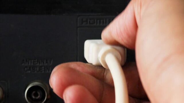 Close-up of a hand plugging a white HDMI cable into an HDMI port on the back of a television.