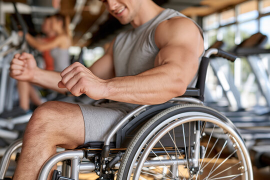 Strong man in wheelchair smiles confidently at gym. Muscular arms flex as he grips his wheel. Background shows blurred exercise equipment and people. Ideal for fitness, disability inclusion