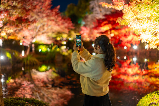 Young female tourist using her smartphone to capture the magical autumn night illumination in a Japanese garden