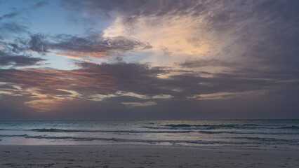 Fototapeta premium A calm evening after sunset.A tropical island.Lilac clouds in the sky are illuminated by golden and crimson light.Waves of the turquoise ocean roll towards the shore, spreading across the sandy beach