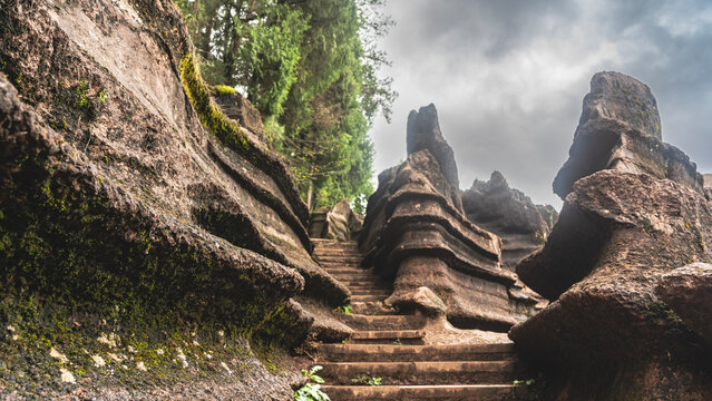A stone staircase rises up a hill between red-brown karst rocks. Steep undulating slopes, with geological layers. Green vegetation. Clouds in the sky. China. Red Stone Forest National Geopark. 

 