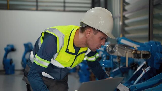 Worker checks robot systems with a laptop. He wears a hard hat and safety vest. Focused, he monitors machines on-site