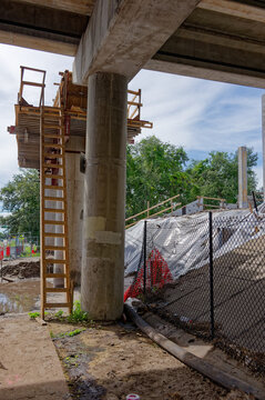 Bridge construction site on a highway overpass featuring scaffolding, concrete pillars, orange traffic barriers and cones, urban infrastructure work and road maintenance under a blue sky with clouds.
