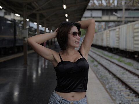 Young Asian woman wearing sunglasses and a black corset top posing with hands behind her head at a train station