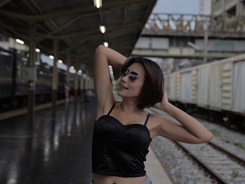 Young Asian woman wearing sunglasses and a black corset top posing with hands behind her head at a train station