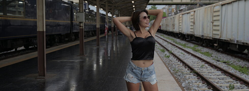 Young Asian woman wearing sunglasses and a black corset top posing with hands behind her head at a train station
