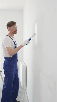 Professional painter wearing protective white gloves and blue work overalls, applying paint with roller on pristine interior wall during home renovation project