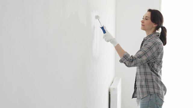Woman wearing protective gloves rolling white paint onto wall, updating living space with fresh color and personal home renovation effort