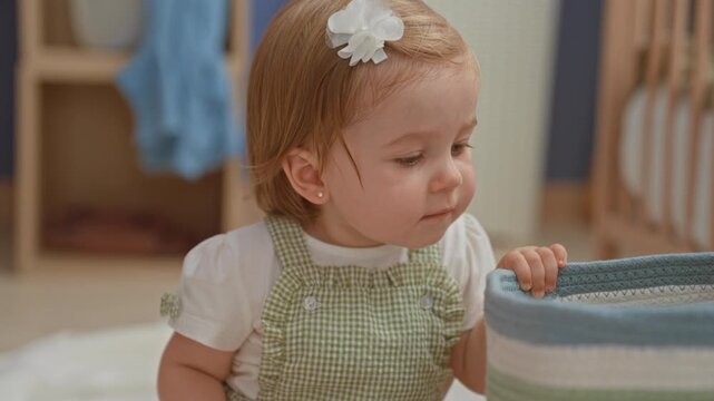 Toddler girl holding pink rattle with small hand, looking up while seated on blanket in building nursery room; curiosity playfulness.