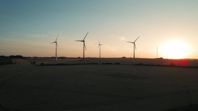 Wind turbines stand tall as the sun sets, illuminating the rolling hills and casting long shadows across the fields.