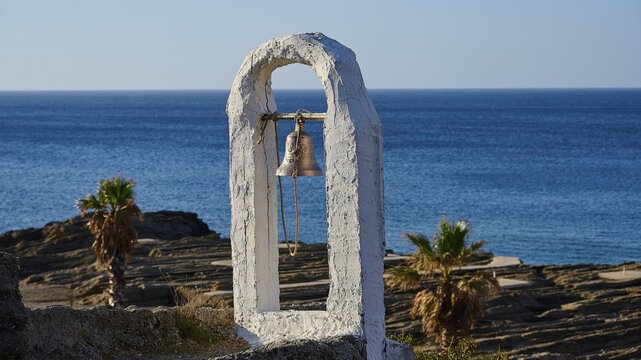 Close-up of a white bell frame on a coastline with palm trees in the background under a sunny blue sky, rock church Agios Nikolaos, Oasis Beach, Kallithea, Rhodes, Dodecanese, Greek Islands, Greece