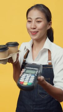 Vertical Screen : Happy smiling Asia waitress wearing apron friendly looking at camera giving coffee or tea and credit card reader machine to customer isolated on yellow background. Entrepreneur cafe.