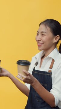 Vertical Screen : Happy Asia lady business owner wearing apron friendly look at advertising space hold coffee or tea isolated on the yellow background. Advertising area, mock up promotional content.