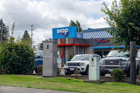 Exterior of an IHOP pancake restaurant with its blue and red logo sign, seen across the street past parked cars and grey utility cabinets on an overcast morning.