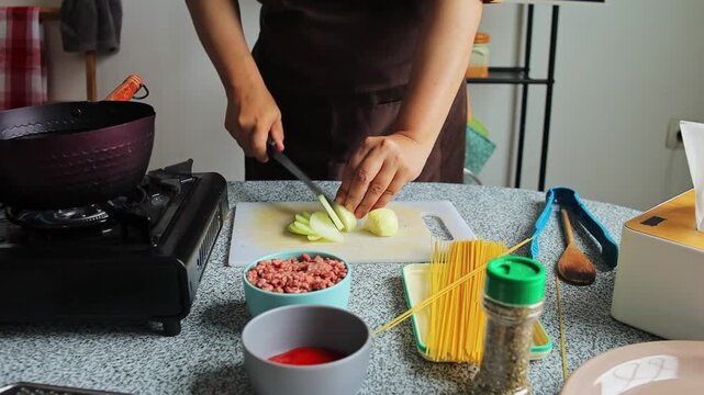 Home cook using sharp knife to slice yellow onion on plastic cutting board for spaghetti preparation