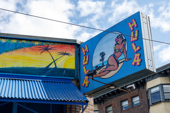 The bright blue Hula Hula tiki bar sign with a painted hula dancer on a surfboard and a sunset palm mural, above a corrugated blue awning near Seattle Center.