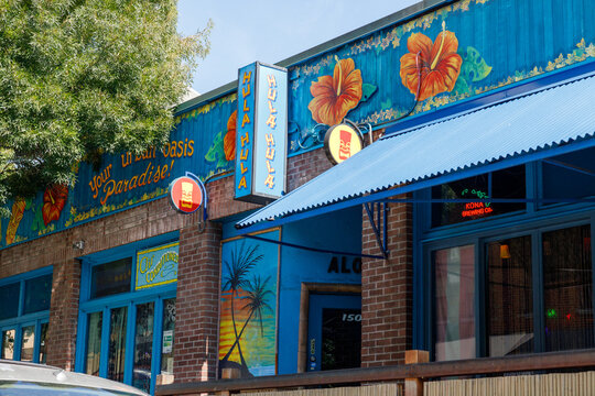The colorful storefront of the Hula Hula tiki bar, tiki mask signs, palm and hibiscus murals and an urban oasis paradise slogan on the blue Lower Queen Anne facade.