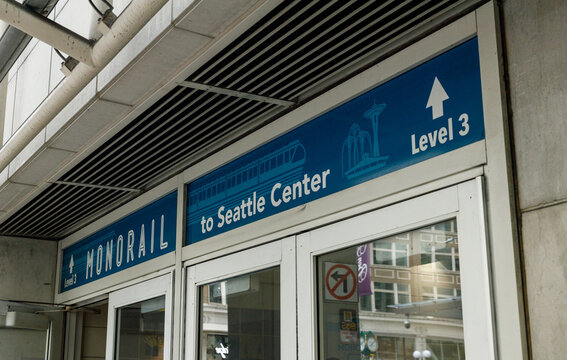 A blue Monorail to Seattle Center Level 3 sign with a train graphic and Space Needle icon, above glass entrance doors at the Westlake Center station downtown.