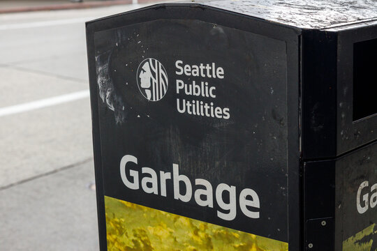 The corner of a black Seattle Public Utilities garbage bin, its white logo and lettering above a worn, sticker marked metal panel on a downtown sidewalk.