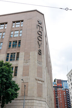 The stone corner of the historic Macy's department store building rises above a carved The Bon Marche nameplate, ornate art deco detailing against an overcast sky downtown.