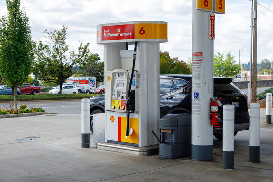 A white Shell V Power Nitro fuel pump numbered 6 stands between white bollards, a black SUV fuelling beside a U Haul truck parked across the lot.