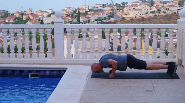 Side profile shot of a mature athletic man performing intense burpees and knuckle push ups on a fitness mat on a villa terrace next to a pool.
