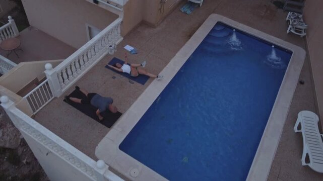 High angle aerial shot of a man doing push ups and a woman doing core leg raises on yoga mats beside a swimming pool at a private villa terrace
