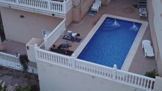 High angle aerial shot of a man doing push ups and a woman doing donkey kicks on yoga mats next to a private villa swimming pool terrace.
